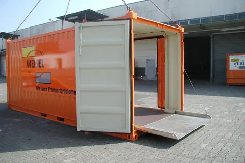 Orangefarbener Weisel-Container mit offener Tür und Laderampe am Kranhaken vor einer Lagerhalle